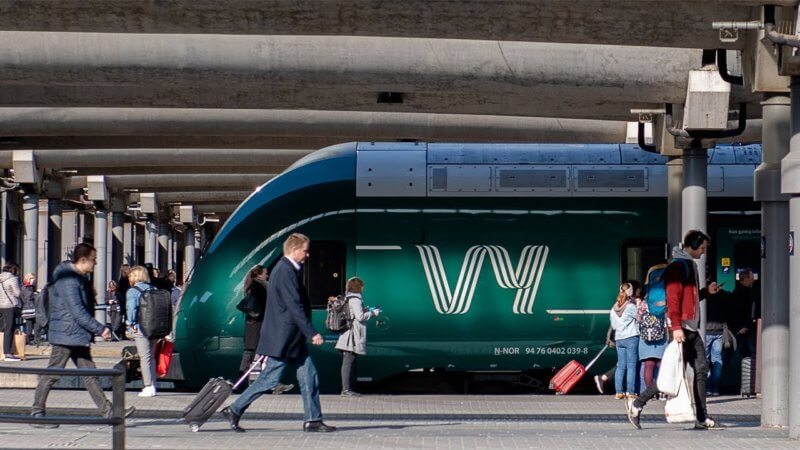 Pendlere travler på en jernbanestasjonsplattform ved siden av et grønt Vy-tog med den hvite logoen CIM 23.02. Noen mennesker går, mens andre dveler i nærheten av det delvis synlige toget under betongtak, og fanger et travelt reisemiljø.